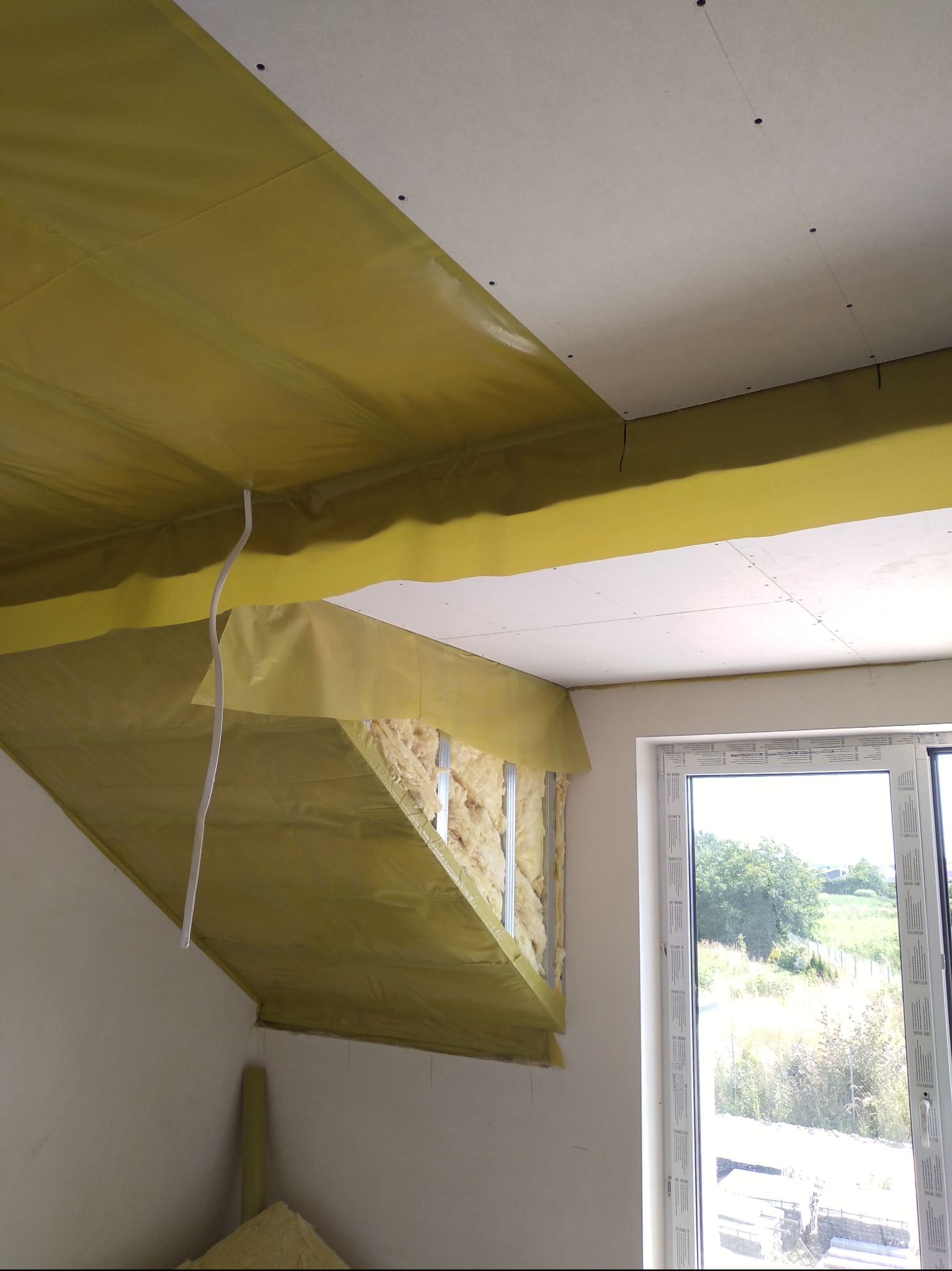 Montaż izolacji z wełny mineralnej i folii paroizolacyjnej na poddaszu, widoczne fragmenty płyty gipsowo-kartonowej na suficie oraz okno z widokiem na zewnątrz.