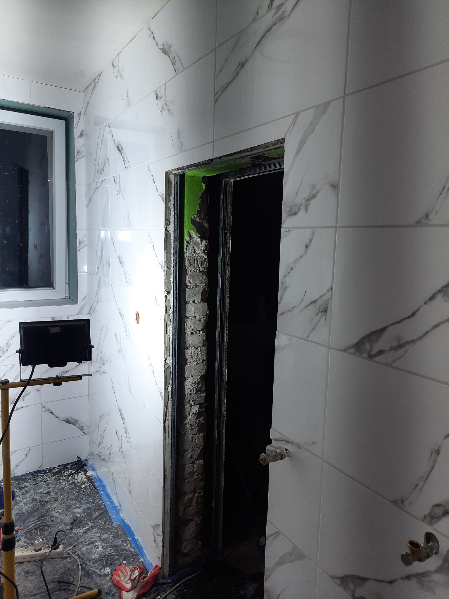 Łazienka w trakcie prac: białe płytki imitujące marmur na ścianach, przygotowane ościeże drzwiowe z odsłoniętym murem, zabezpieczona folią podłoga, lampa robocza oświetlająca wnętrze.