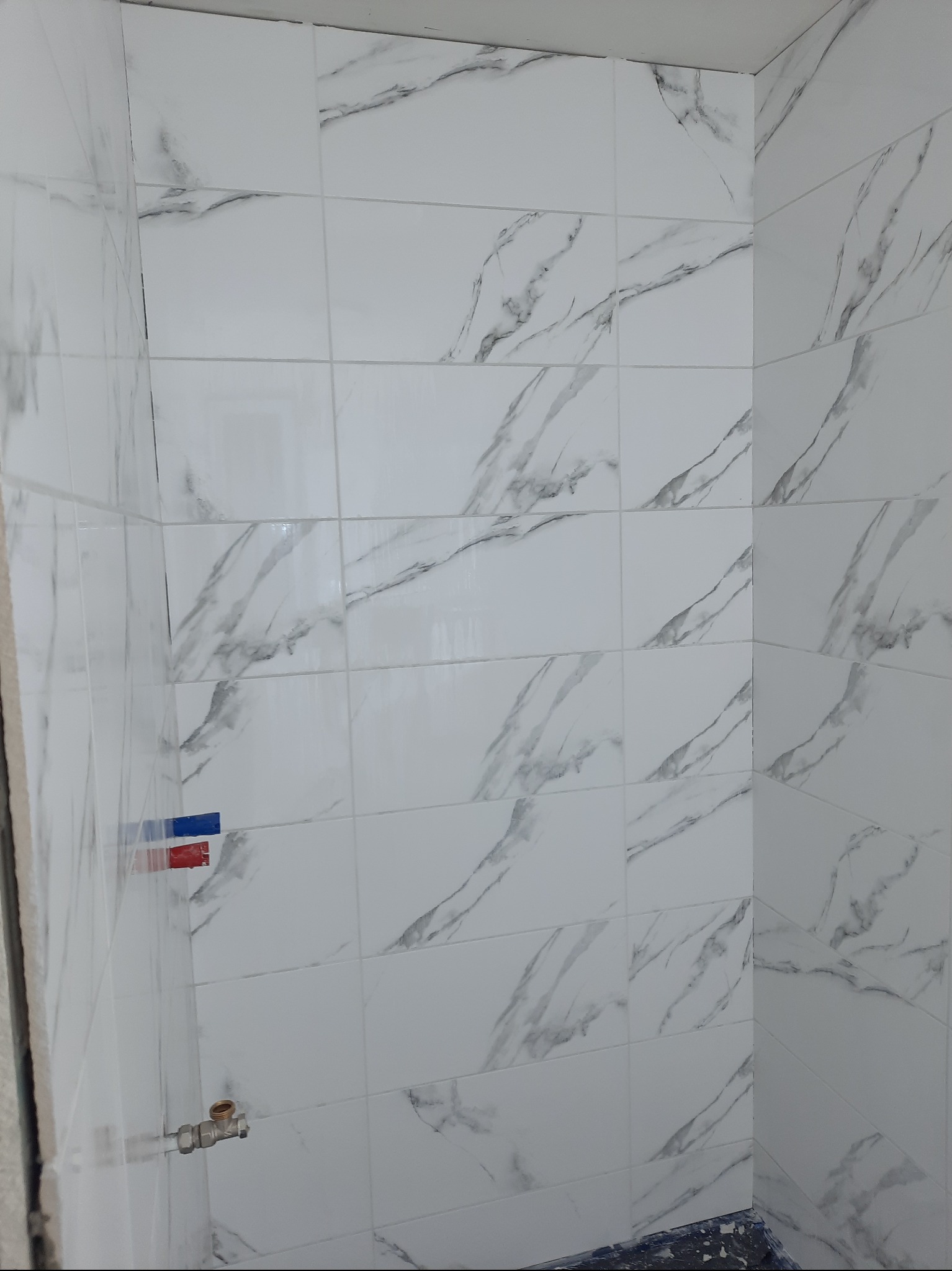 Ściana łazienki wyłożona białymi płytkami imitującymi marmur, widoczne przyłącze wodne i fragment zabezpieczonej podłogi.