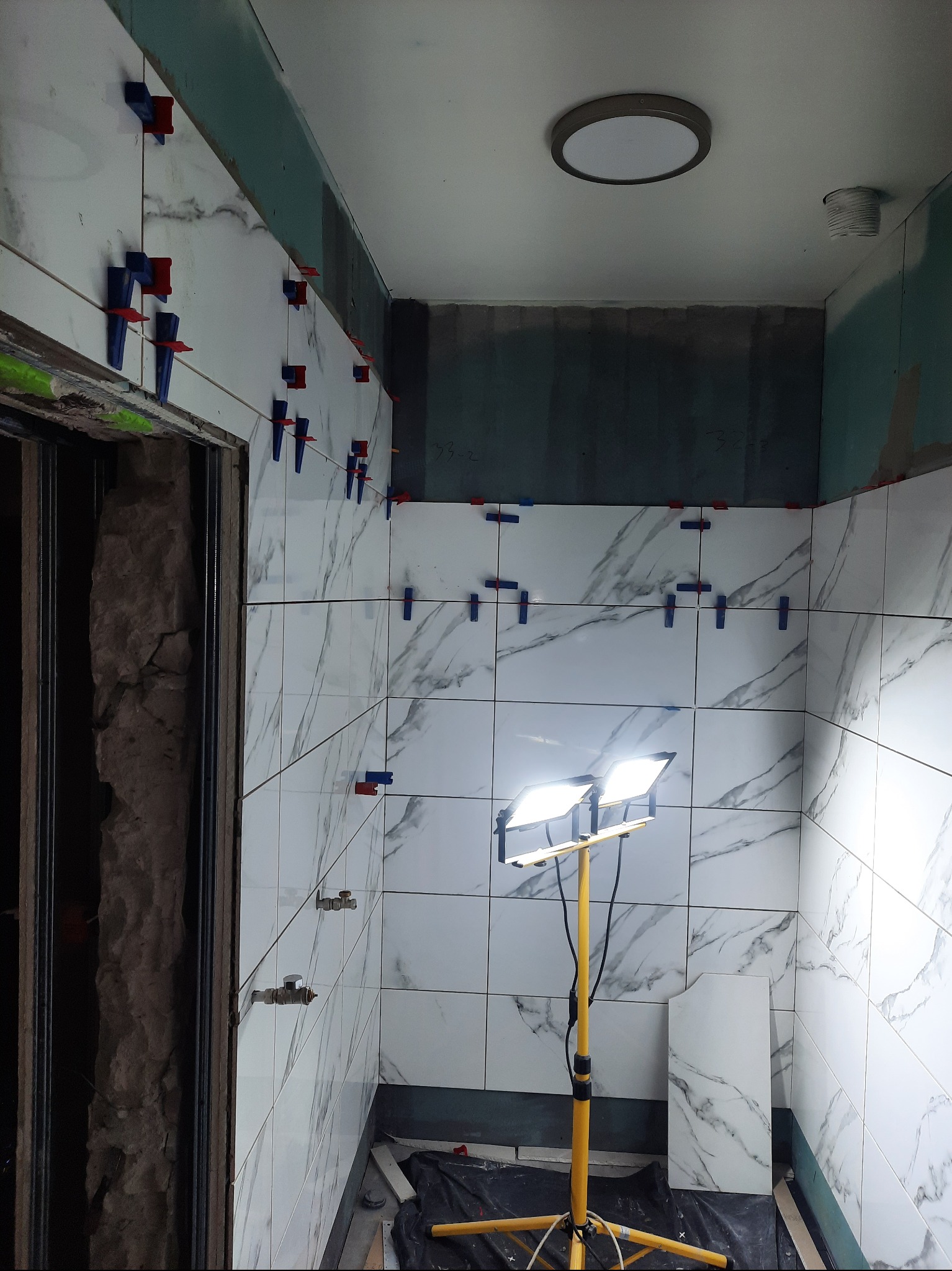 Montaż płytek marmuropodobnych w łazience, widoczne krzyżyki dystansowe, oświetlenie robocze na statywie i fragment ściany przygotowanej pod płytki.