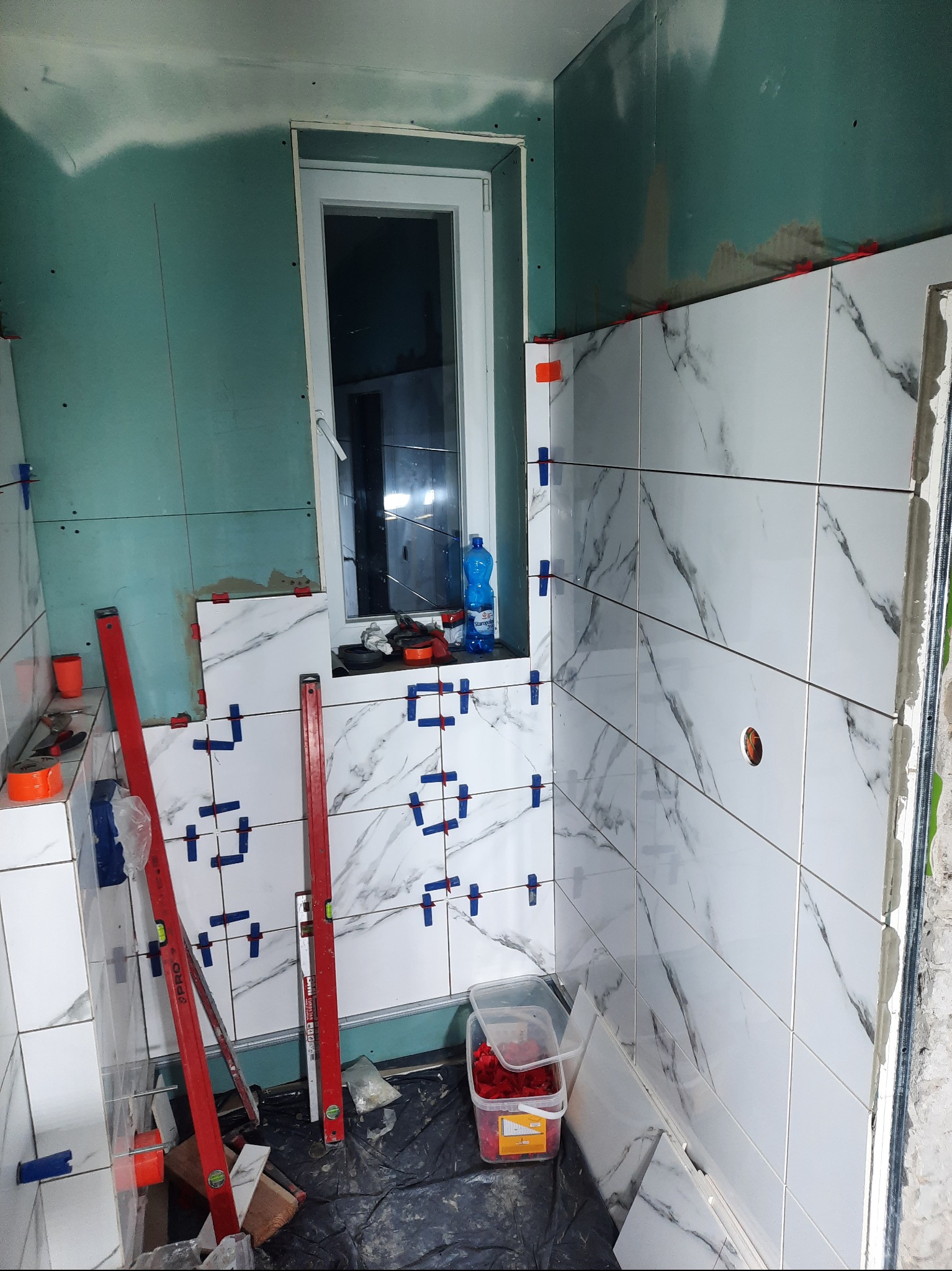 Wnętrze łazienki w trakcie układania białych płytek imitujących marmur, widoczne poziomice, plastikowe krzyżyki dystansowe, niezabudowana ściana z płyty gipsowo-kartonowej oraz okno.