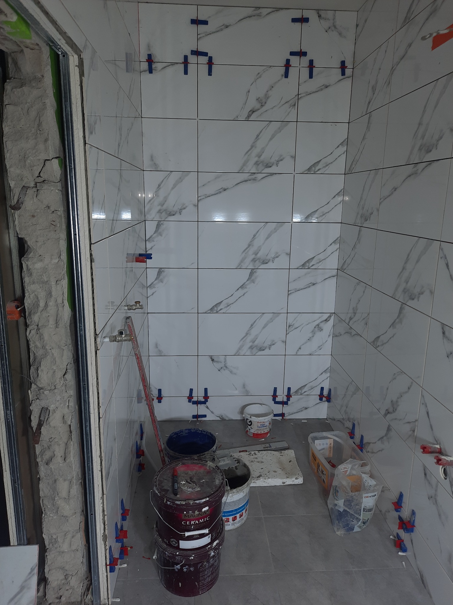 Wnętrze łazienki w trakcie układania białych płytek imitujących marmur, widoczne plastikowe krzyżyki dystansowe, wiaderka z zaprawą, poziomica oraz surowy fragment ściany przy futrynie.