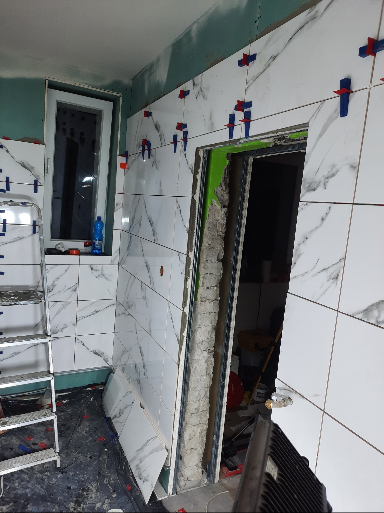 Białe płytki imitujące marmur, ułożone na ścianie łazienki w trakcie prac, z użyciem klipsów poziomujących; widoczna drabina, okno i surowa ściana z płyty gipsowo-kartonowej.