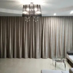 Długie, beżowe zasłony zakrywające całą ścianę, wiszące pod sufitem, oświetlone przez czarny żyrandol z kryształkami, widok z wnętrza salonu.