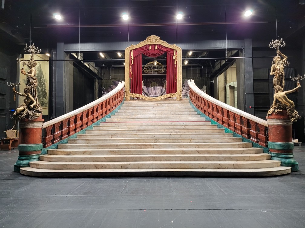 Scena teatralna z marmurowymi schodami, balustradą w kolorze bordo, złotymi rzeźbami z kandelabrami i dekoracyjnym złotym portalem z czerwonymi zasłonami w tle, oświetlona reflektorami.