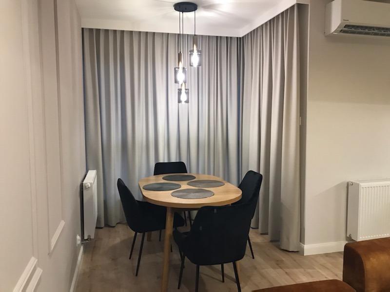 Jadalnia z owalnym stołem z jasnego drewna, czarnymi krzesłami, szarymi zasłonami od podłogi do sufitu i wiszącą lampą z trzema szklanymi kloszami.