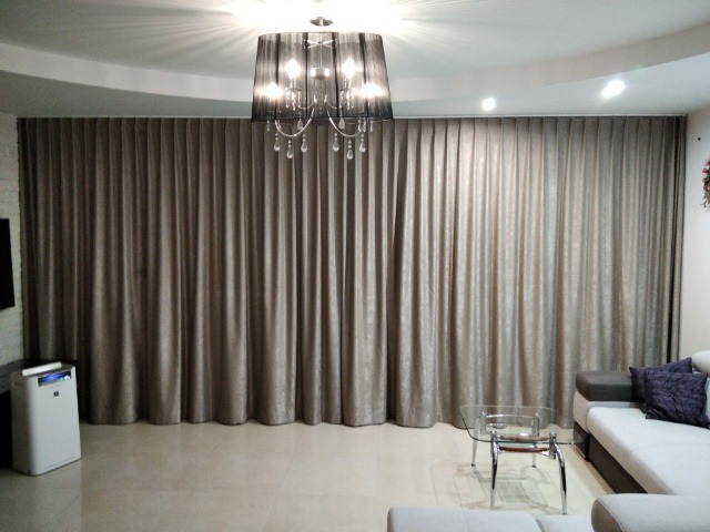 Długie, beżowe zasłony zakrywające całą ścianę, wiszące pod sufitem, oświetlone przez czarny żyrandol z kryształkami, widok z wnętrza salonu.