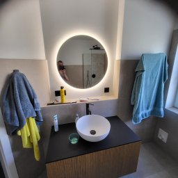 Adam Janduda - Nowoczesna łazienka z okrągłym lustrem LED, białą umywalką nablatową na szafce z lamelami i szarymi płytkami. Widoczne akcesoria łazienkowe i odbicie osoby w lustrze.