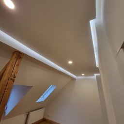 Adam Janduda - Wykończone poddasze w Rudzie Śląskiej: drewniana podłoga, okno dachowe i nowoczesne oświetlenie sufitowe z listwami LED. Widok z dołu, podkreślający skosy i detale wykończenia.