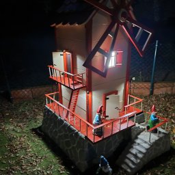 Adam Janduda - Oświetlony miniaturowy wiatrak z czerwonymi akcentami i figurkami gnomów w ogrodzie nocą. Dekoracja z kamienną podstawą i schodami.