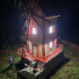 Adam Janduda - Oświetlony, miniaturowy, dwupiętrowy wiatrak z czerwonymi akcentami, otoczony figurkami krasnali ogrodowych na trawniku w nocy. Widoczne detale konstrukcyjne i oświetlenie okien.