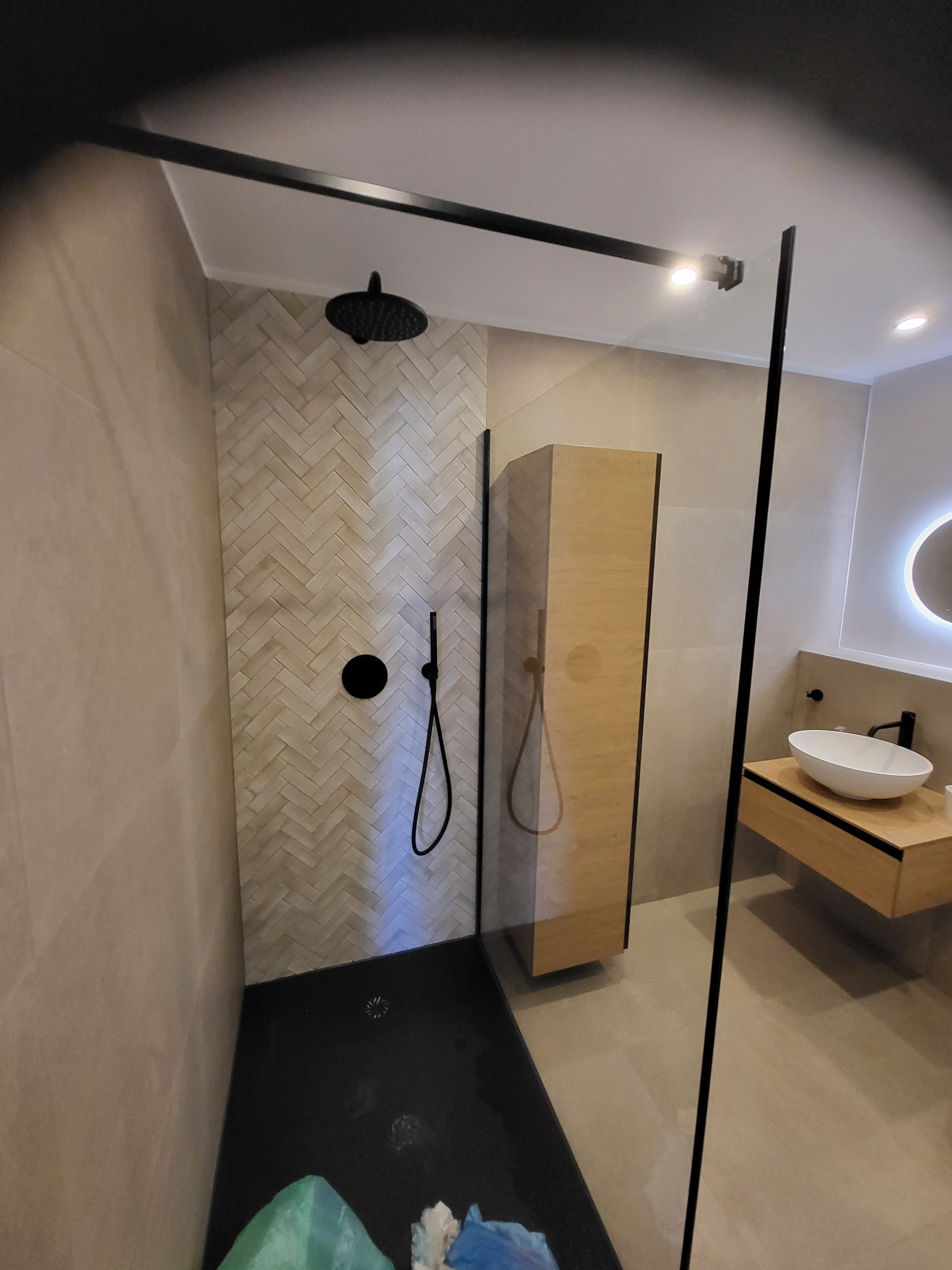 Nowoczesna łazienka z kabiną typu walk-in, czarną armaturą i drewnianą szafką pod umywalką. Ściana wyłożona płytkami w jodełkę. Minimalistyczny design.