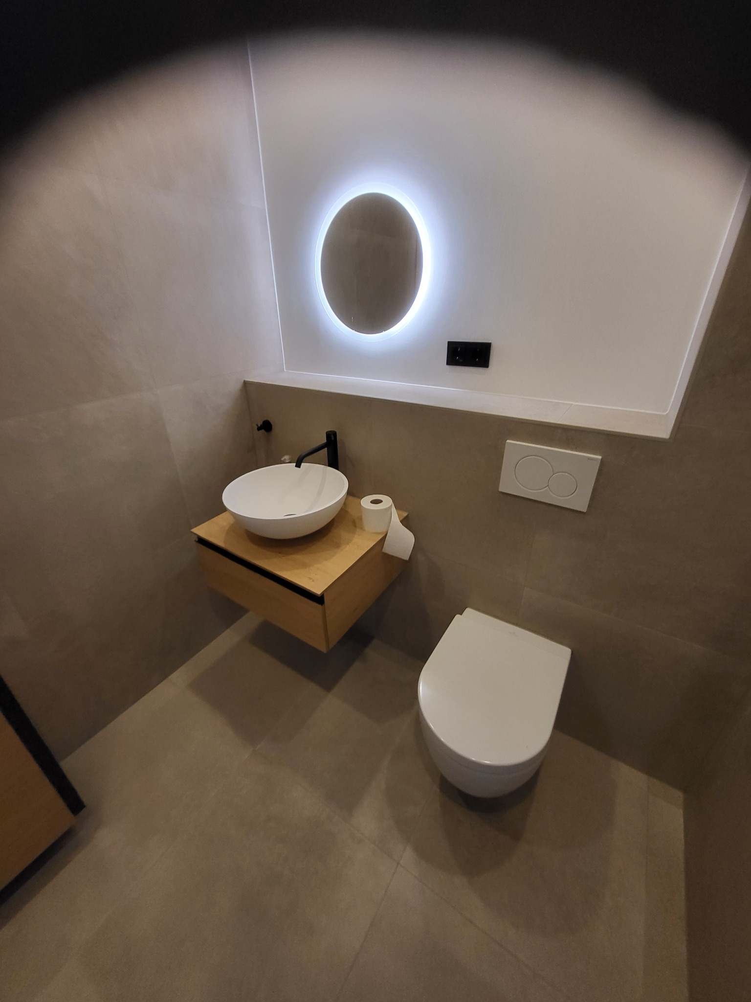 Nowoczesna łazienka z okrągłym lustrem podświetlanym LED, umywalką nablatową na drewnianej szafce i podwieszaną toaletą. Stonowana kolorystyka płytek.