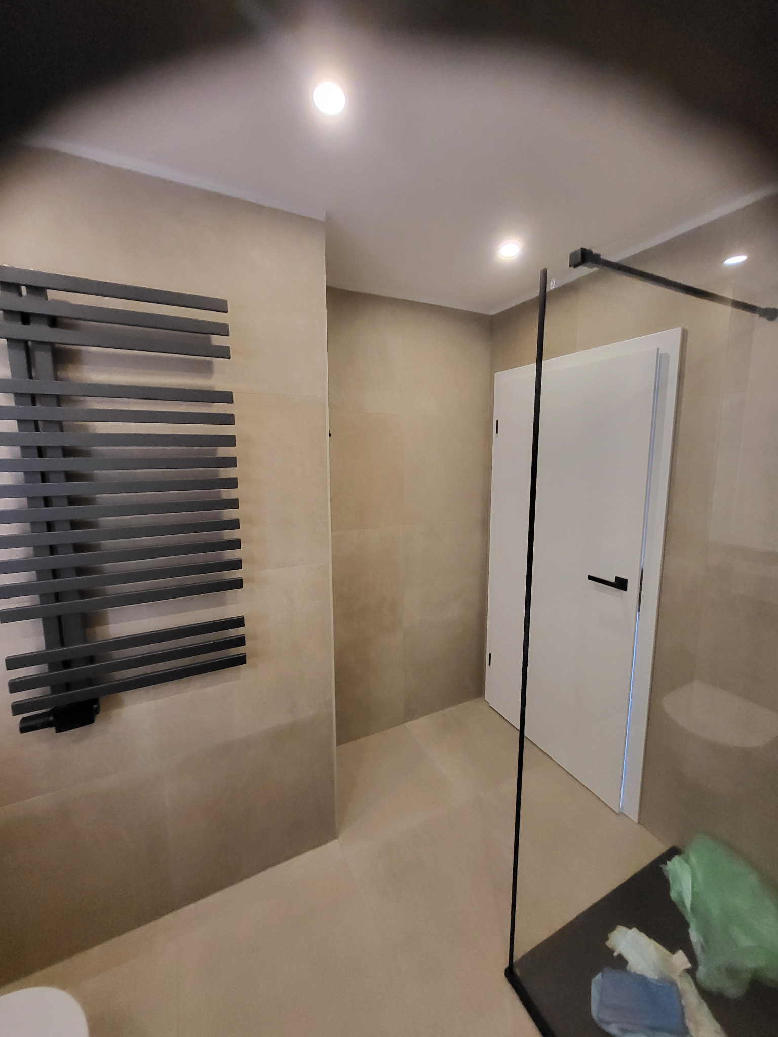 Beżowa łazienka z grzejnikiem drabinkowym, kabiną prysznicową ze szkła i białą, minimalistyczną armaturą. Widoczne płytki w jednolitym odcieniu i oświetlenie sufitowe.