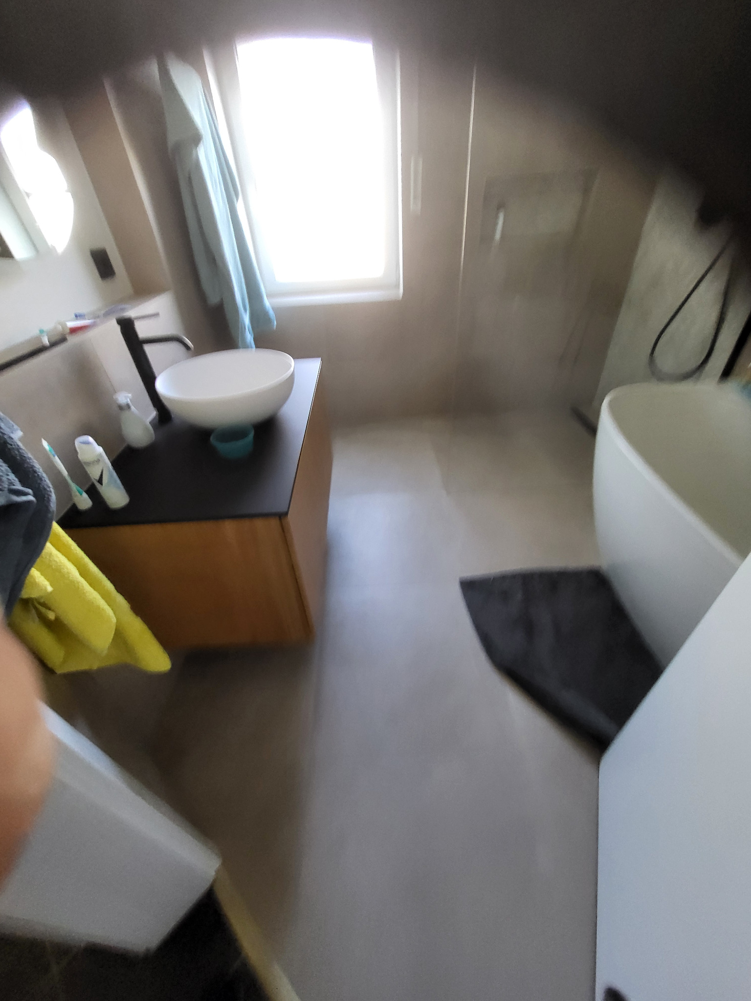 Nowoczesna łazienka z umywalką nablatową, minimalistyczną armaturą i wolnostojącą wanną. Jasne wnętrze z szarymi płytkami i naturalnym światłem z okna.