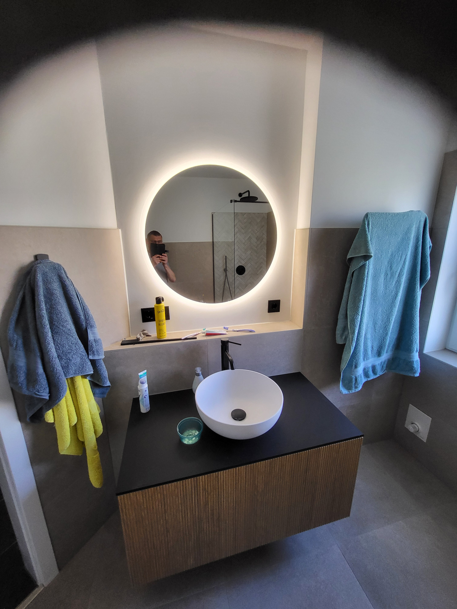 Nowoczesna łazienka z okrągłym lustrem LED, białą umywalką nablatową na szafce z lamelami i szarymi płytkami. Widoczne akcesoria łazienkowe i odbicie osoby w lustrze.