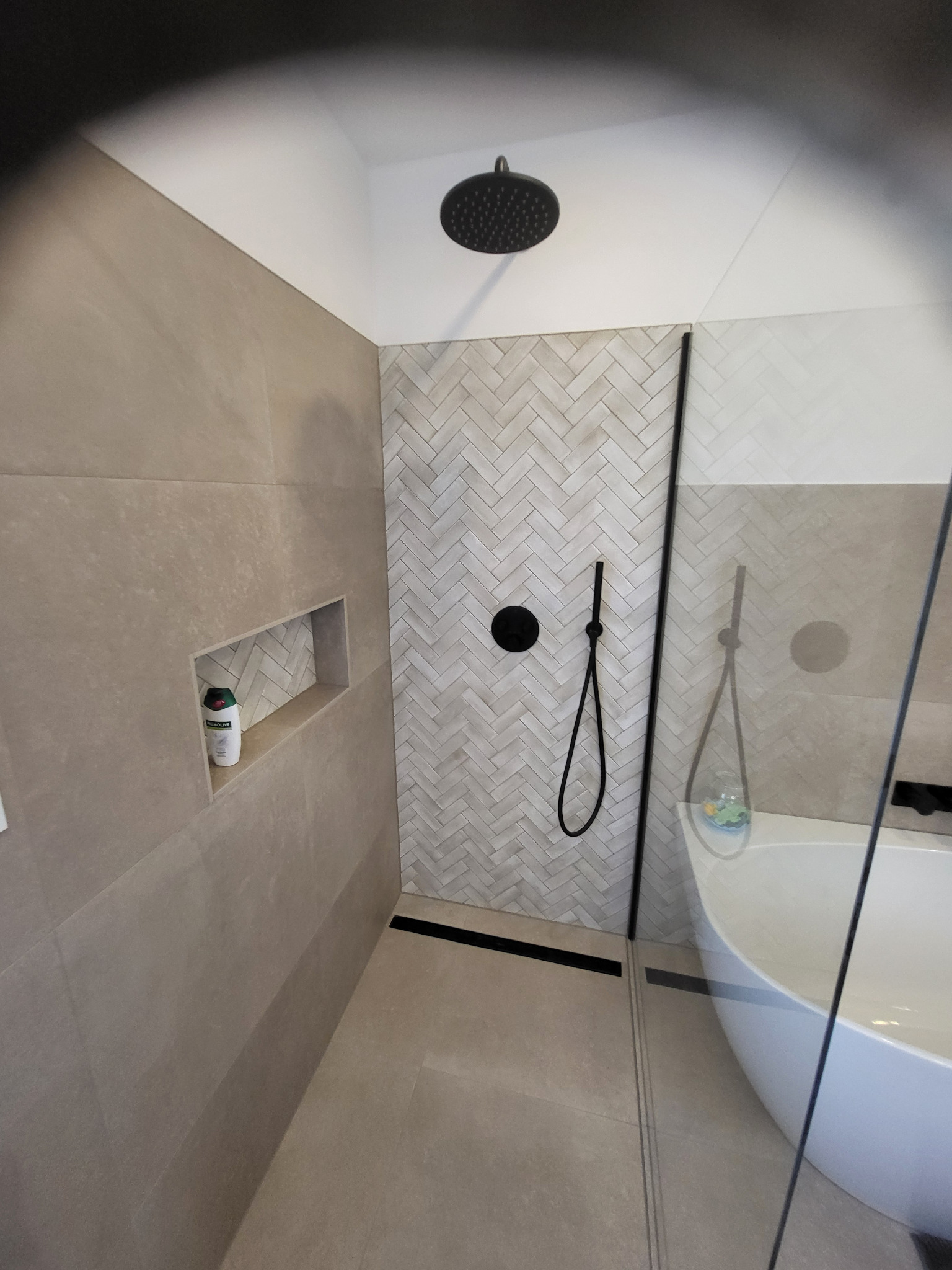 Nowoczesna łazienka z beżowymi płytkami, kabiną prysznicową typu walk-in i czarną armaturą. W ścianie wnęka z kosmetykiem. Ułożenie płytek w jodełkę.