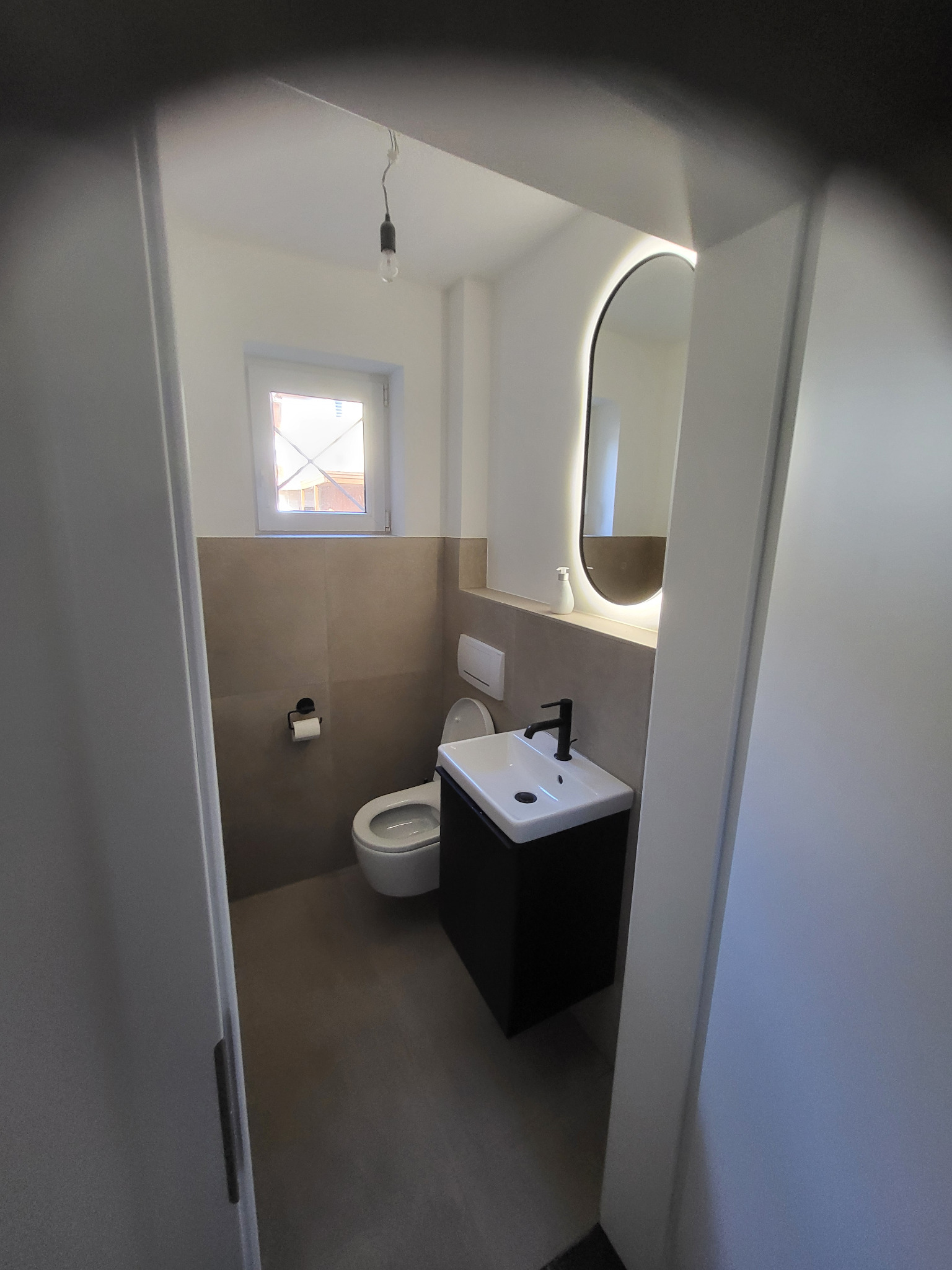 Nowoczesna łazienka z podświetlanym lustrem, umywalką na szafce i toaletą. Ściany wykończone płytkami w dwóch odcieniach beżu. Widok zza uchylonych drzwi.