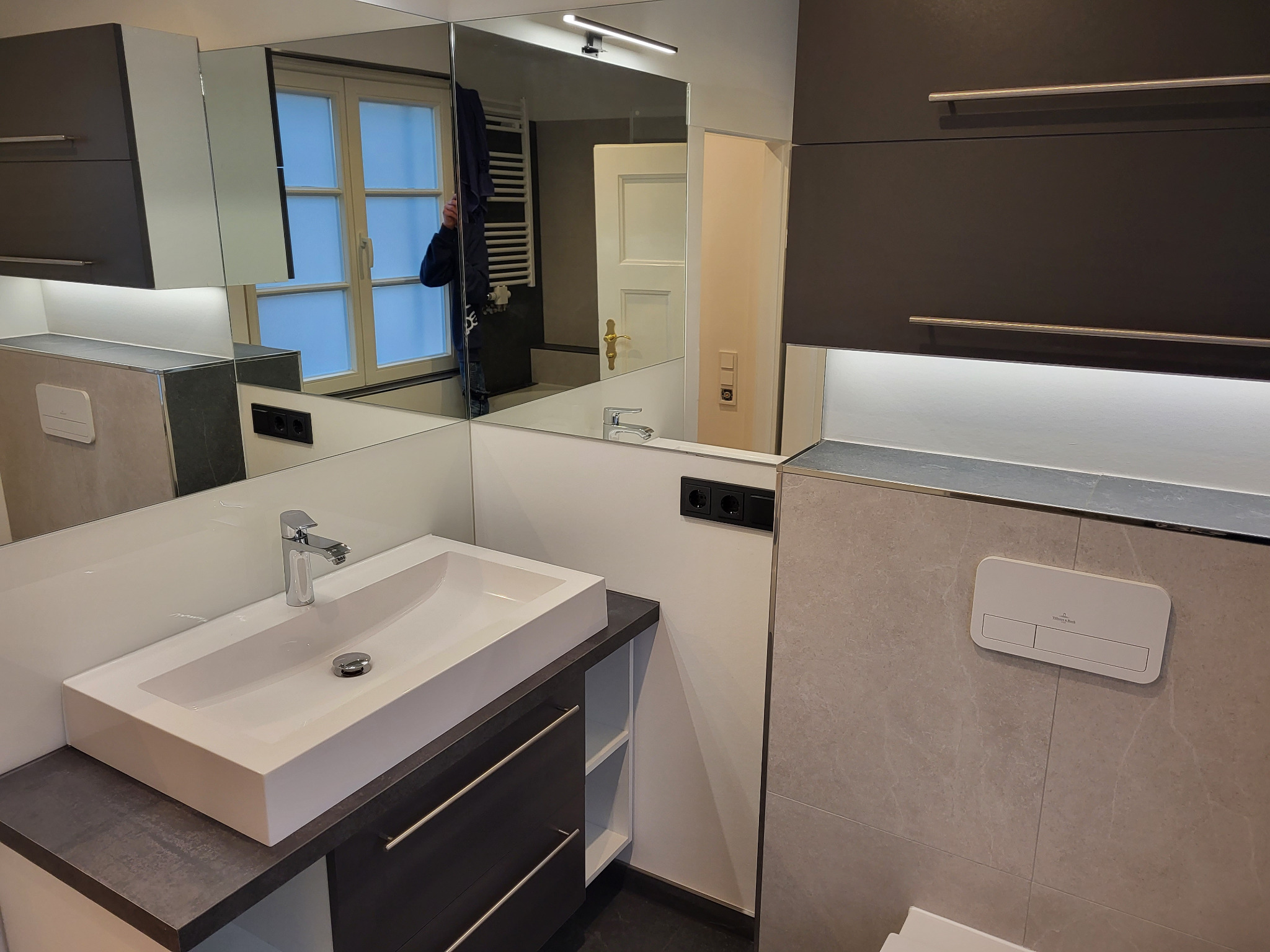 Nowoczesna łazienka z umywalką nablatową, szarą szafką z szufladami i dużym lustrem. Widoczne płytki imitujące marmur i minimalistyczna armatura.