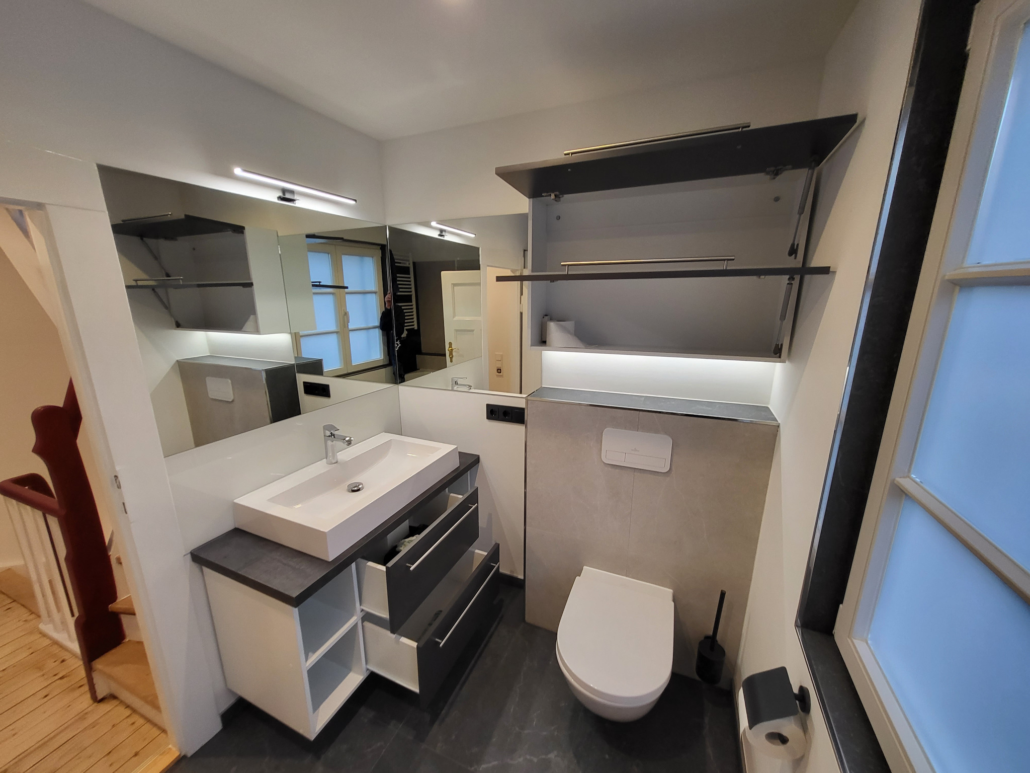 Nowoczesna łazienka po remoncie: biała umywalka z szufladami, podwieszana toaleta, szara płytka, lustro z oświetleniem LED i szafka nad toaletą. Widok z góry.