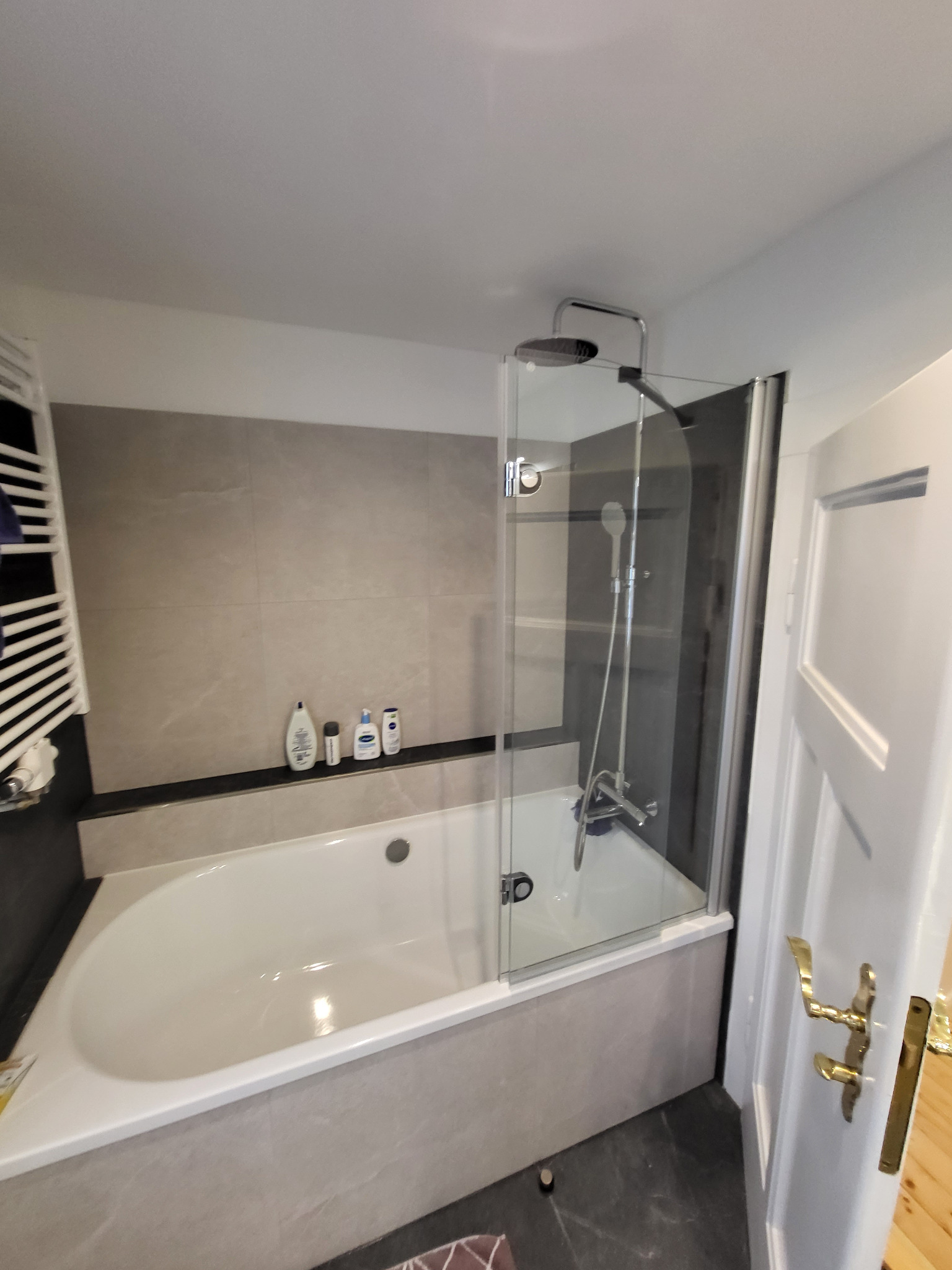 Wykończona łazienka z wanną i szklanym parawanem nawannowym. Na ścianie szare płytki imitujące kamień, biała wanna i grzejnik drabinkowy. Widoczna armatura.