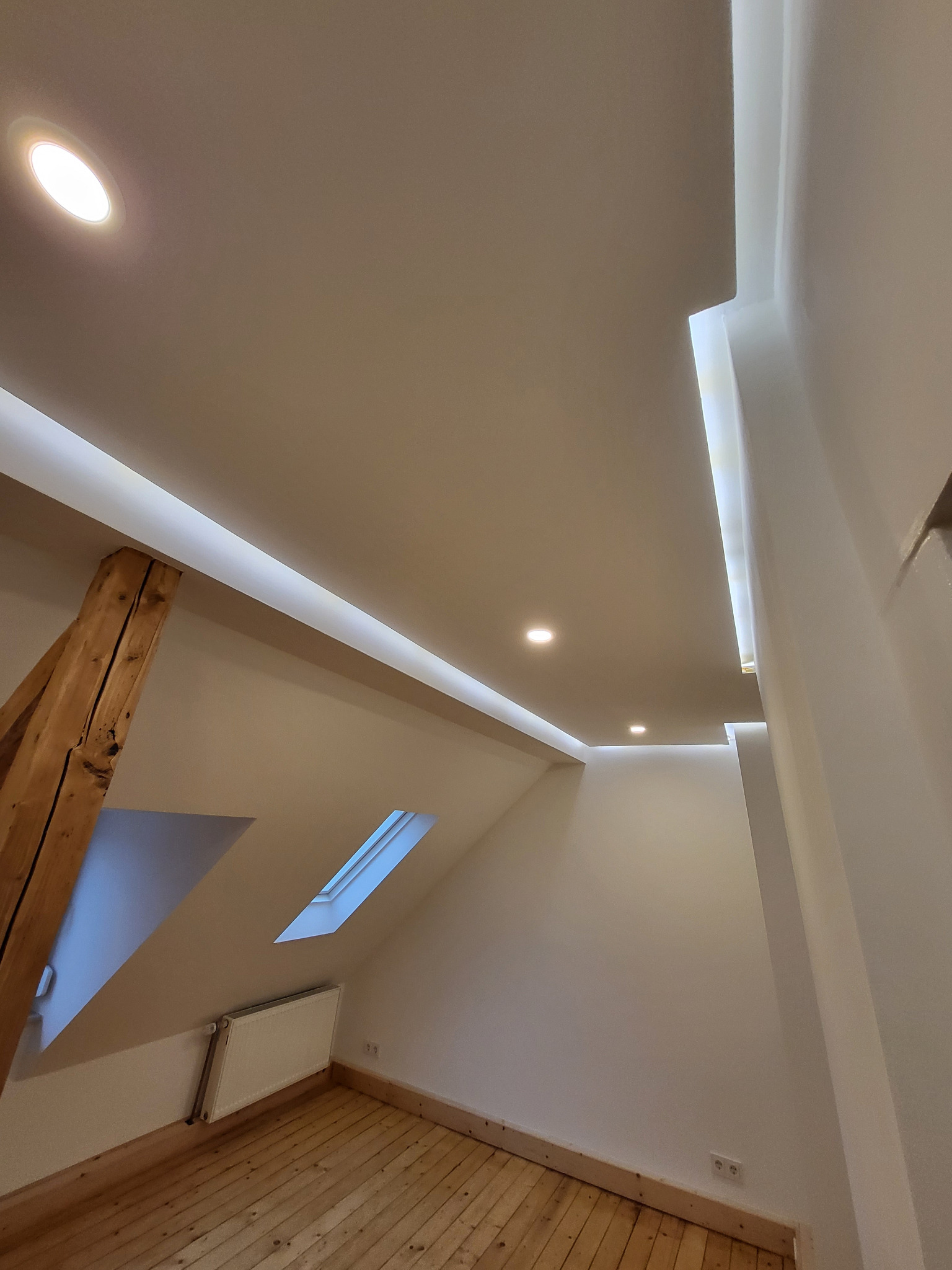 Wykończone poddasze w Rudzie Śląskiej: drewniana podłoga, okno dachowe i nowoczesne oświetlenie sufitowe z listwami LED. Widok z dołu, podkreślający skosy i detale wykończenia.