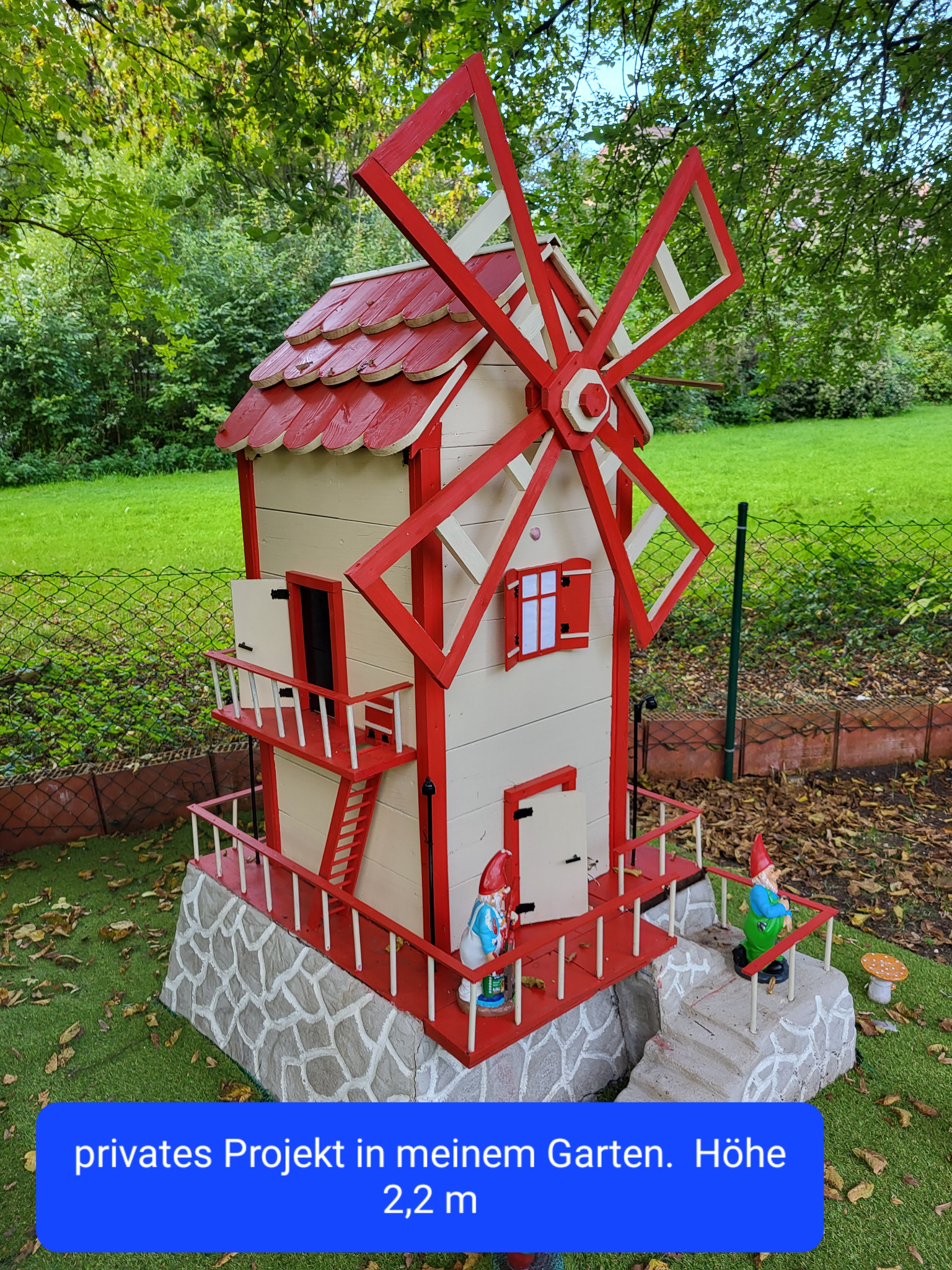 Ogród z dekoracyjnym, drewnianym młynem w kolorach czerwieni i bieli, otoczonym figurkami krasnali i grzybków. Tło stanowi zielona trawa i drzewa.