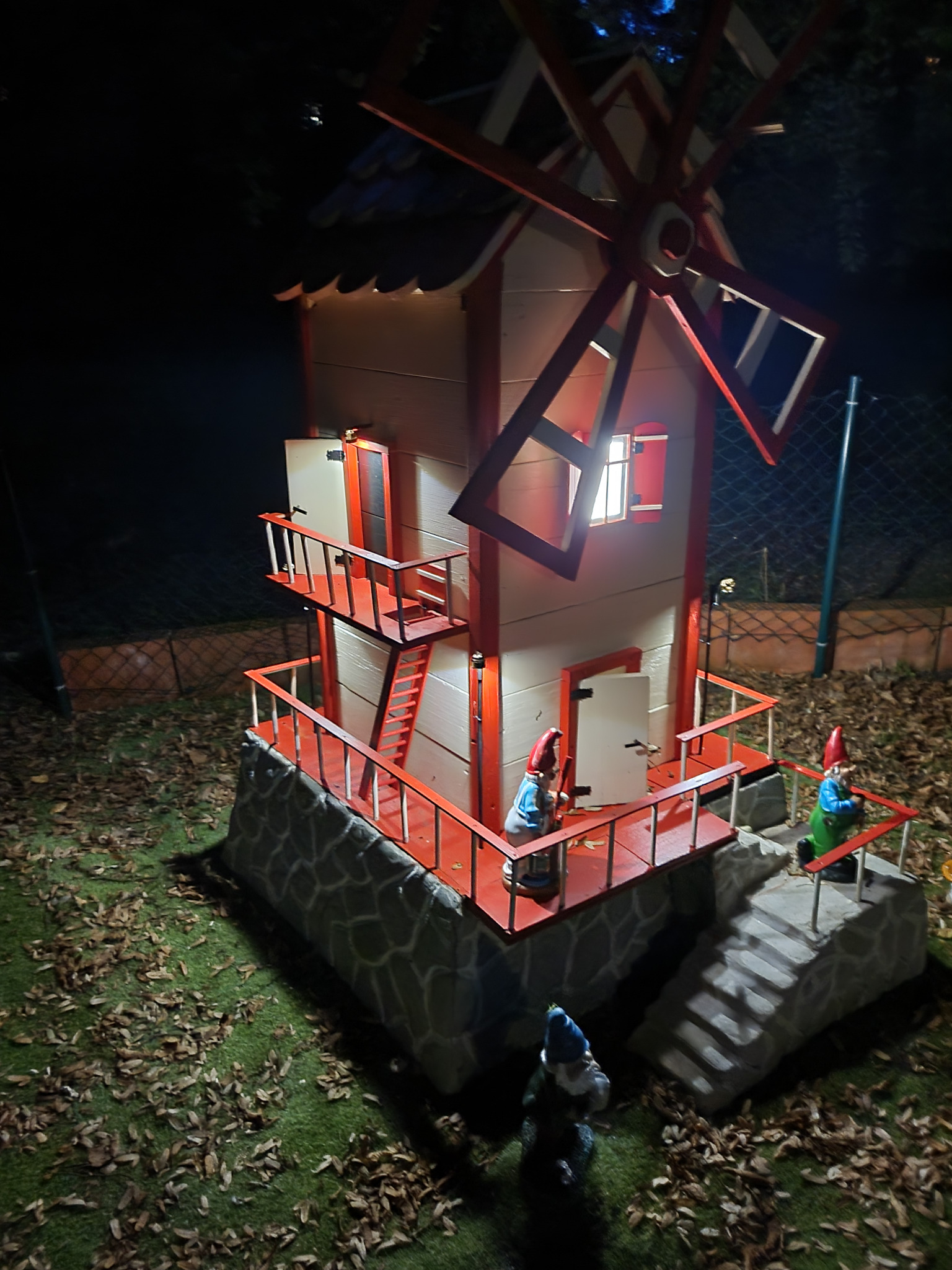 Oświetlony miniaturowy wiatrak z czerwonymi akcentami i figurkami gnomów w ogrodzie nocą. Dekoracja z kamienną podstawą i schodami.