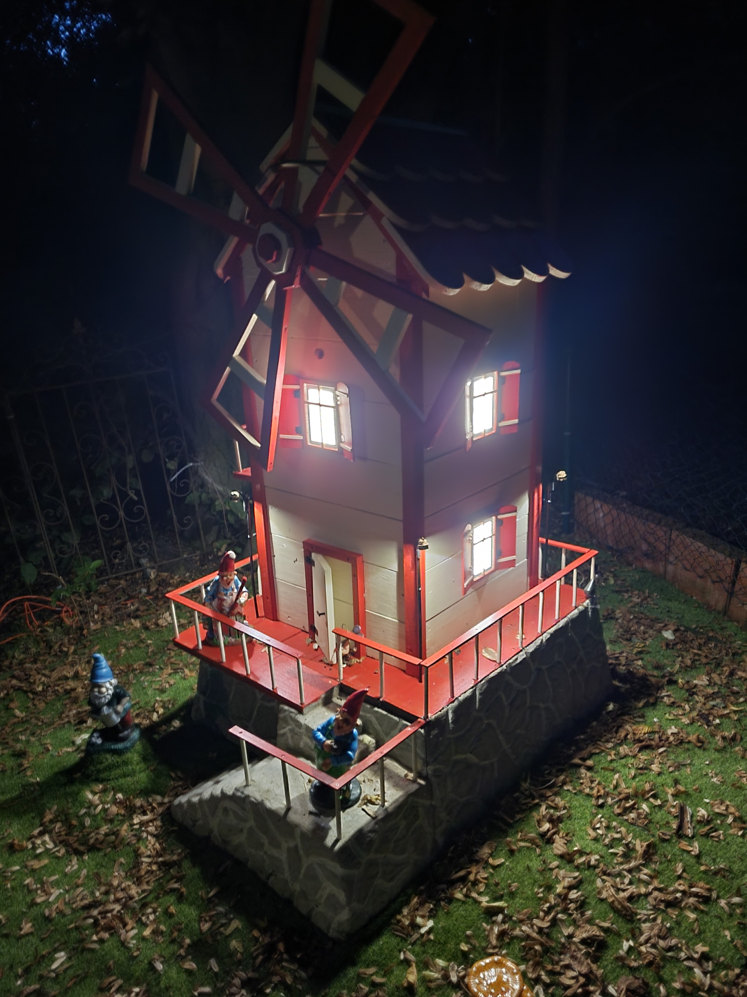 Oświetlony, miniaturowy, dwupiętrowy wiatrak z czerwonymi akcentami, otoczony figurkami krasnali ogrodowych na trawniku w nocy. Widoczne detale konstrukcyjne i oświetlenie okien.