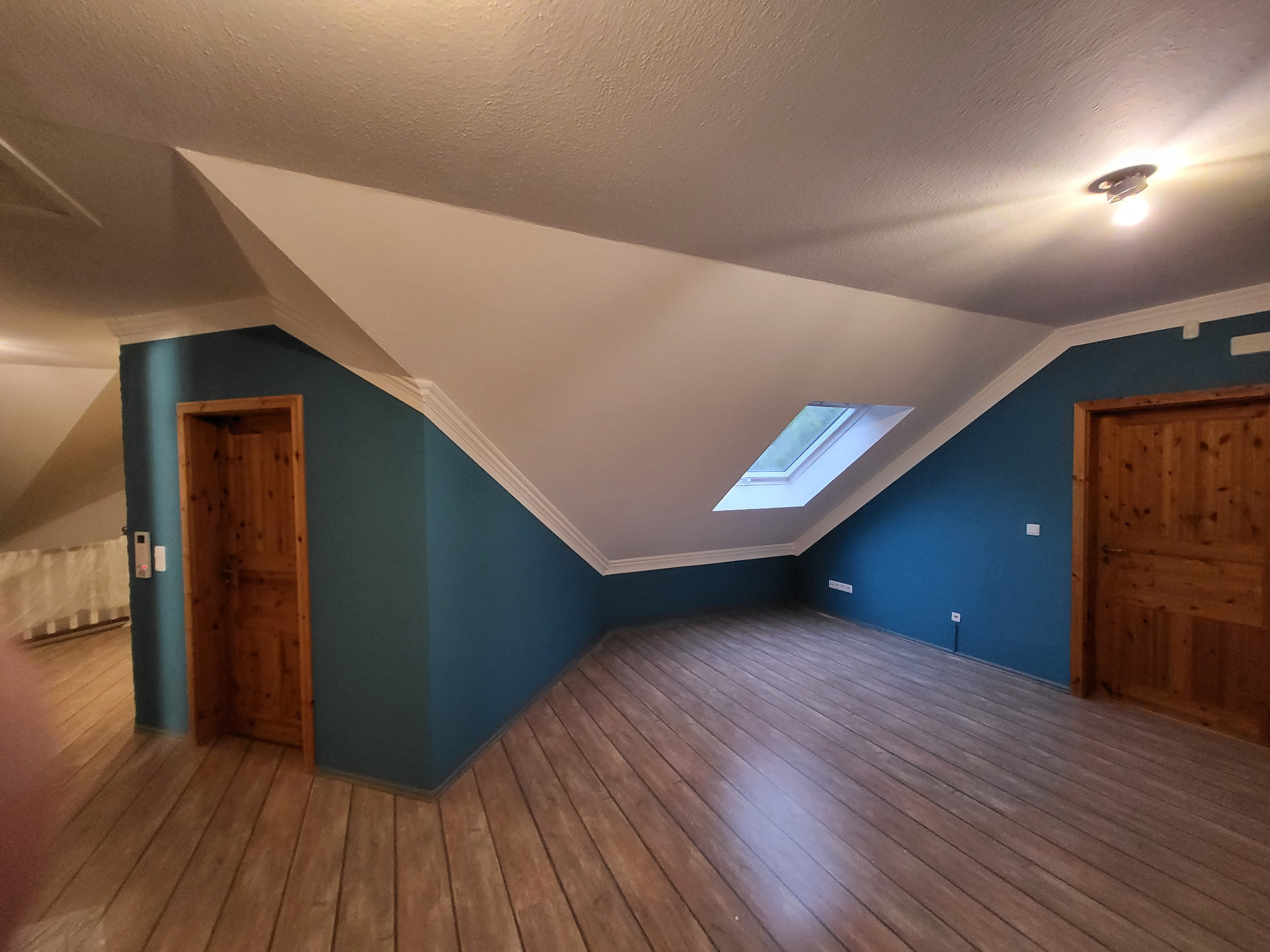 Pomieszczenie na poddaszu po malowaniu ścian na kolor morski, z drewnianą podłogą i oknem dachowym. Widoczne drewniane drzwi i białe wykończenia sufitu.