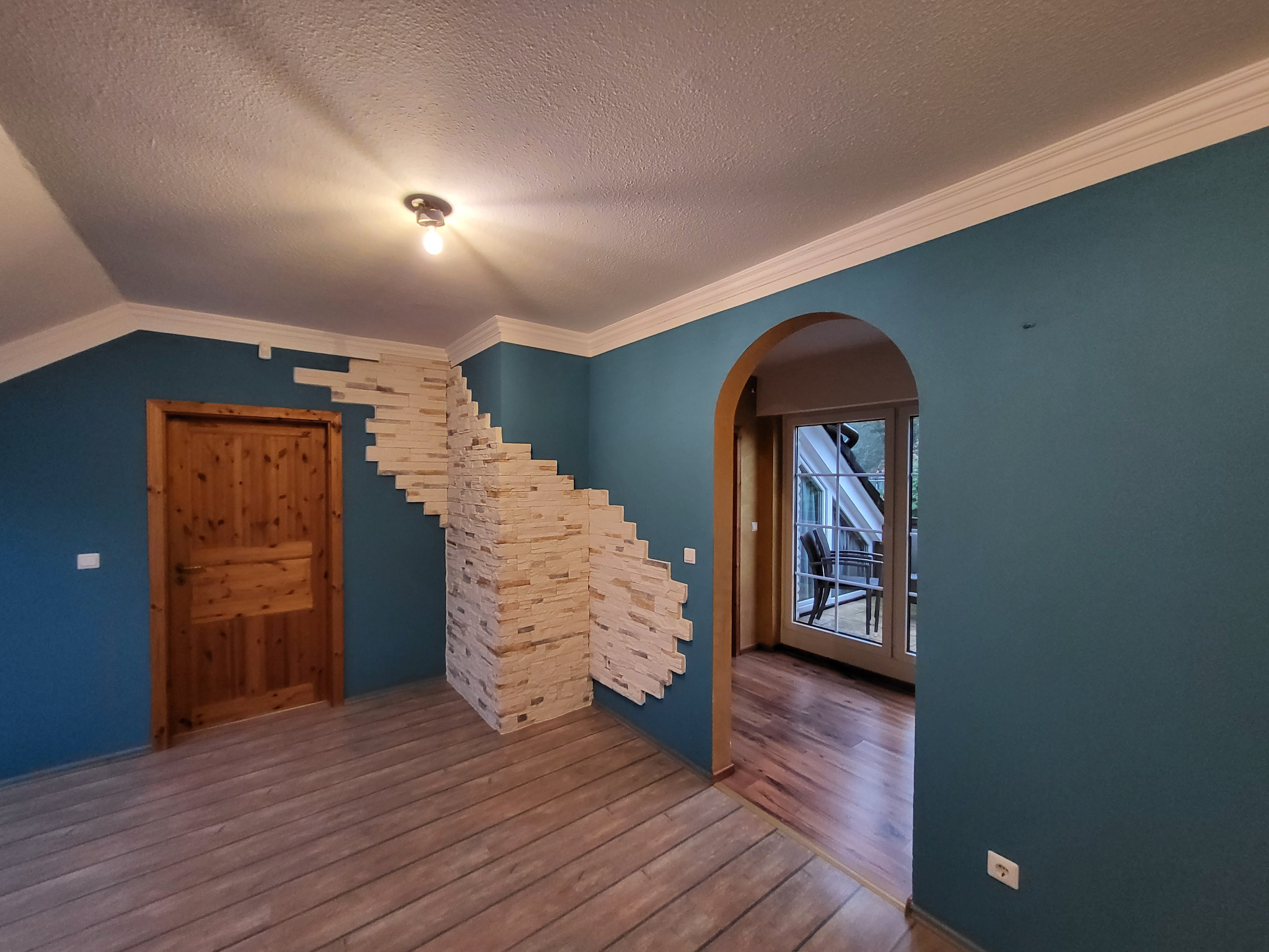 Turkusowe ściany z dekoracyjną, kamienną mozaiką w przestronnym wnętrzu z drewnianą podłogą i drzwiami. Widoczny łuk prowadzący do kolejnego pomieszczenia.