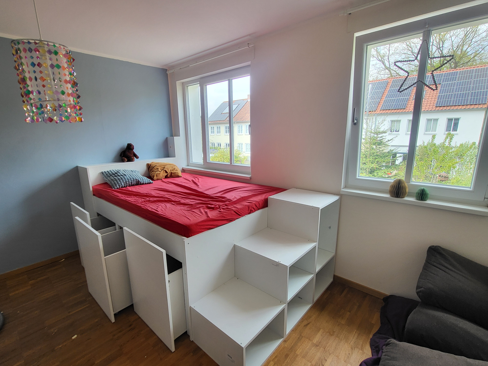 Pokój dziecięcy z łóżkiem na podeście z szufladami i schodkami, czerwona pościel, okno z widokiem na dom z panelami słonecznymi, kolorowa lampa.