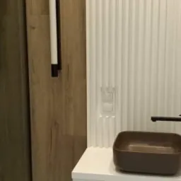 Nowoczesna łazienka z umywalką w kolorze brązu, białą szafką i oświetleniem ściennym z czarnymi detalami na tle ściany z pionowymi żłobieniami.