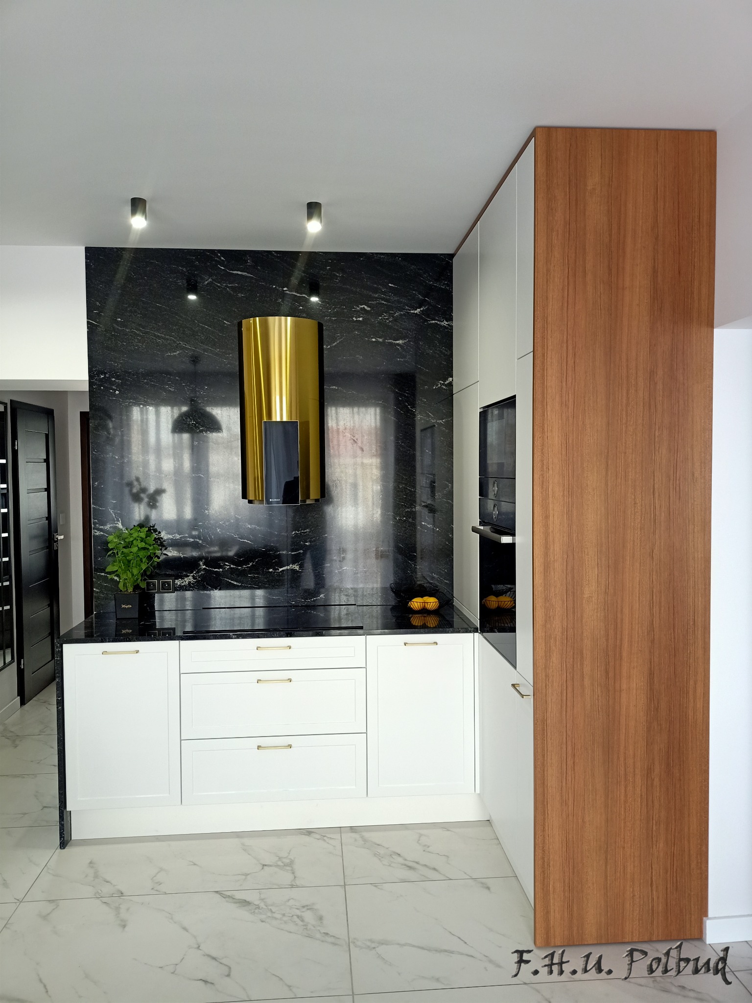 Nowoczesna kuchnia z białymi szafkami, czarnym blatem i złotym okapem cylindrycznym na tle ściany z czarnego marmuru z białymi żyłami, uzupełniona drewnianym panelem bocznym i marmurową podłogą.