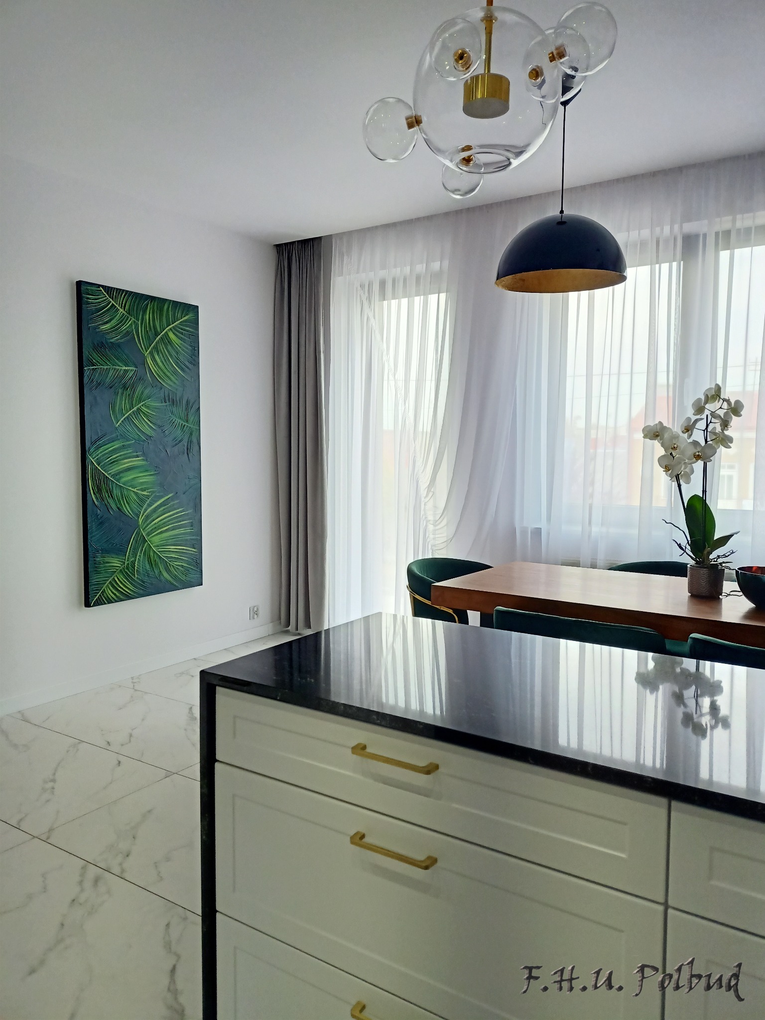 Nowoczesne wnętrze jadalni z marmurową podłogą i wyspą kuchenną z czarnym blatem, obraz z motywem liści na ścianie, okno z białymi zasłonami i designerskie lampy wiszące.