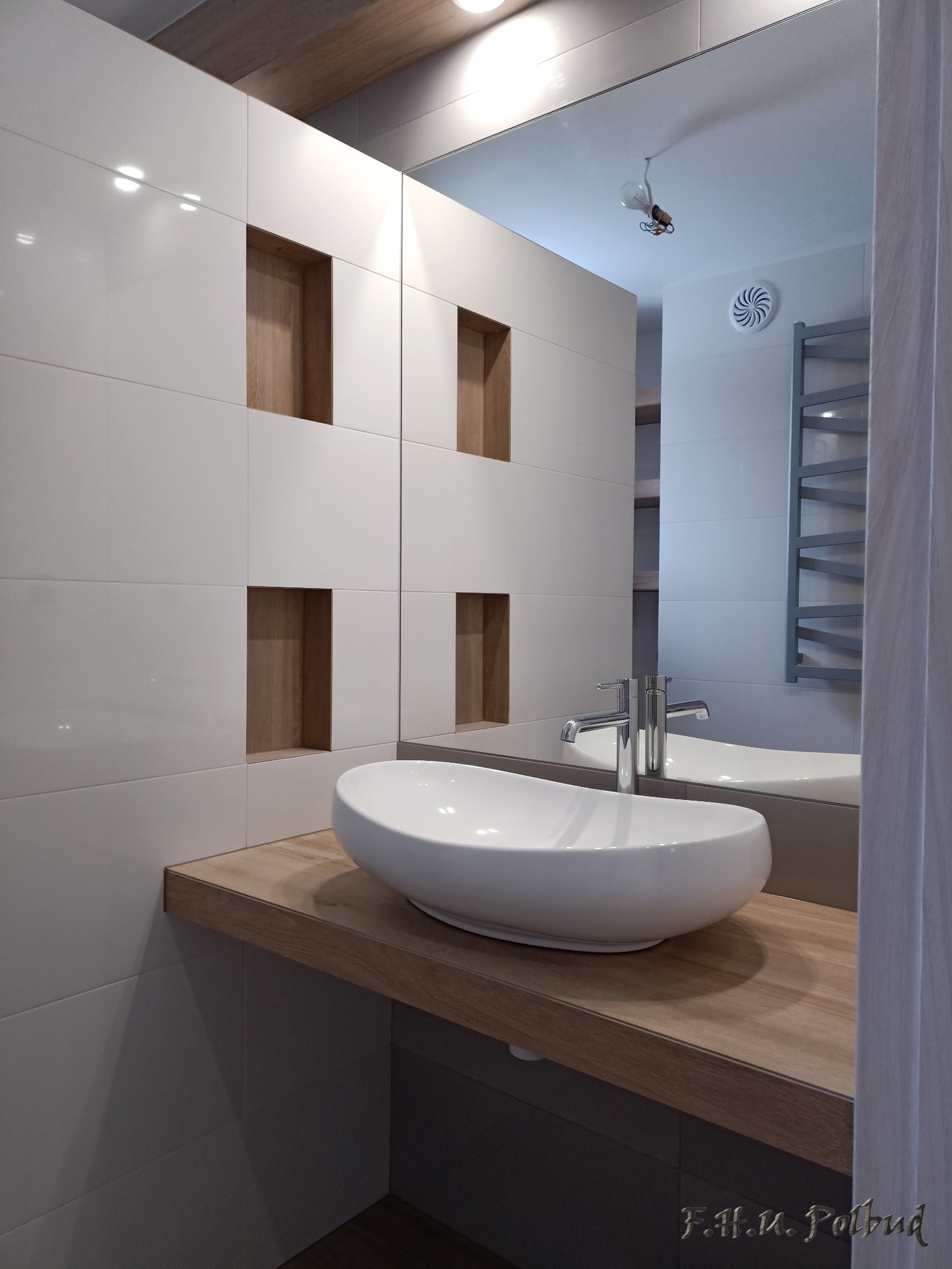 Biała, owalna umywalka nablatowa na drewnianym blacie w nowoczesnej łazience z białymi, błyszczącymi płytkami i wnękami wykończonymi drewnem.