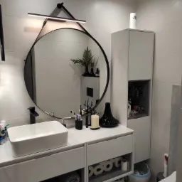 Nowoczesna, jasna łazienka z okrągłym lustrem z podświetleniem LED, białą umywalką nablatową na szafce z szufladami, wysoką szafką z półkami i dekoracjami, oraz elementami wyposażenia jak wazon...