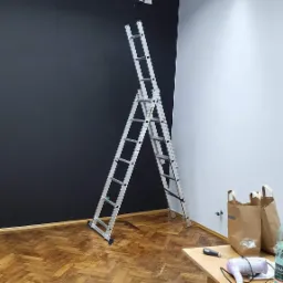 Pokój biurowy z pomalowaną na czarno ścianą, aluminiowa drabina rozstawiona na parkiecie, na biurku suszarka, śrubokręt i butelka wody.