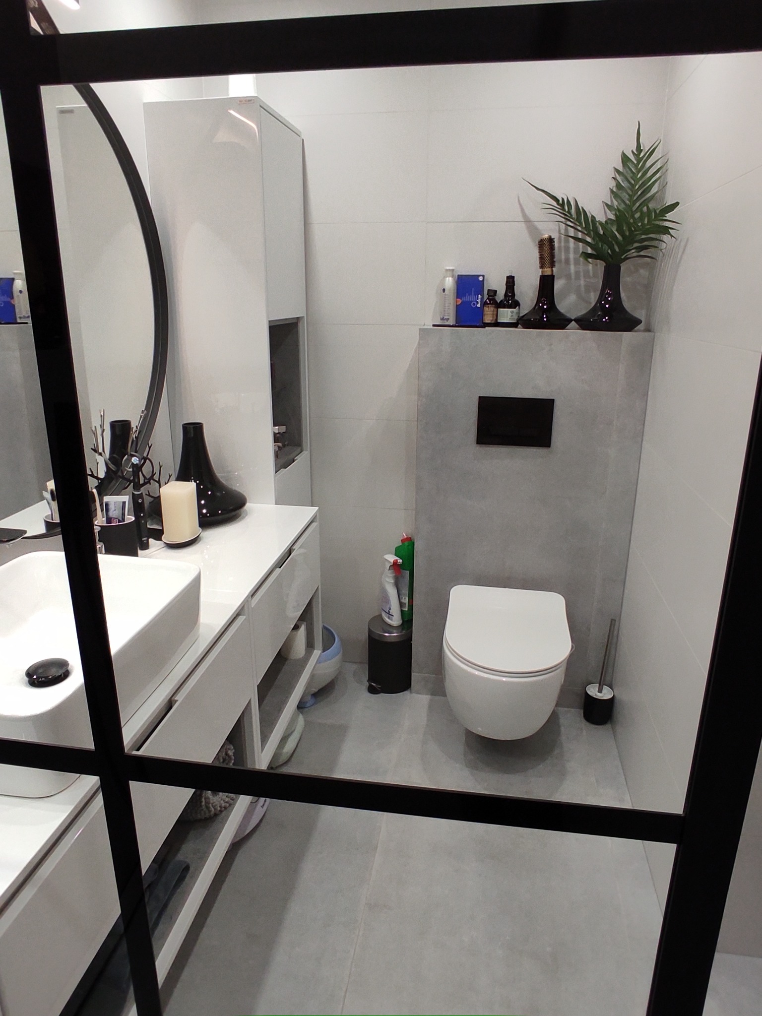 Nowoczesna łazienka z białą umywalką nablatową, toaletą wiszącą i szarymi płytkami, widok przez czarną ramę kabiny prysznicowej.