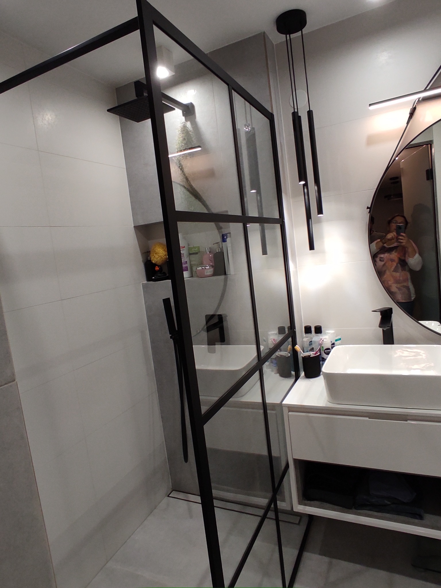 Nowoczesna łazienka z kabiną prysznicową typu walk-in z czarnymi profilami, białą umywalką nablatową i owalnym lustrem z odbiciem osoby robiącej zdjęcie.
