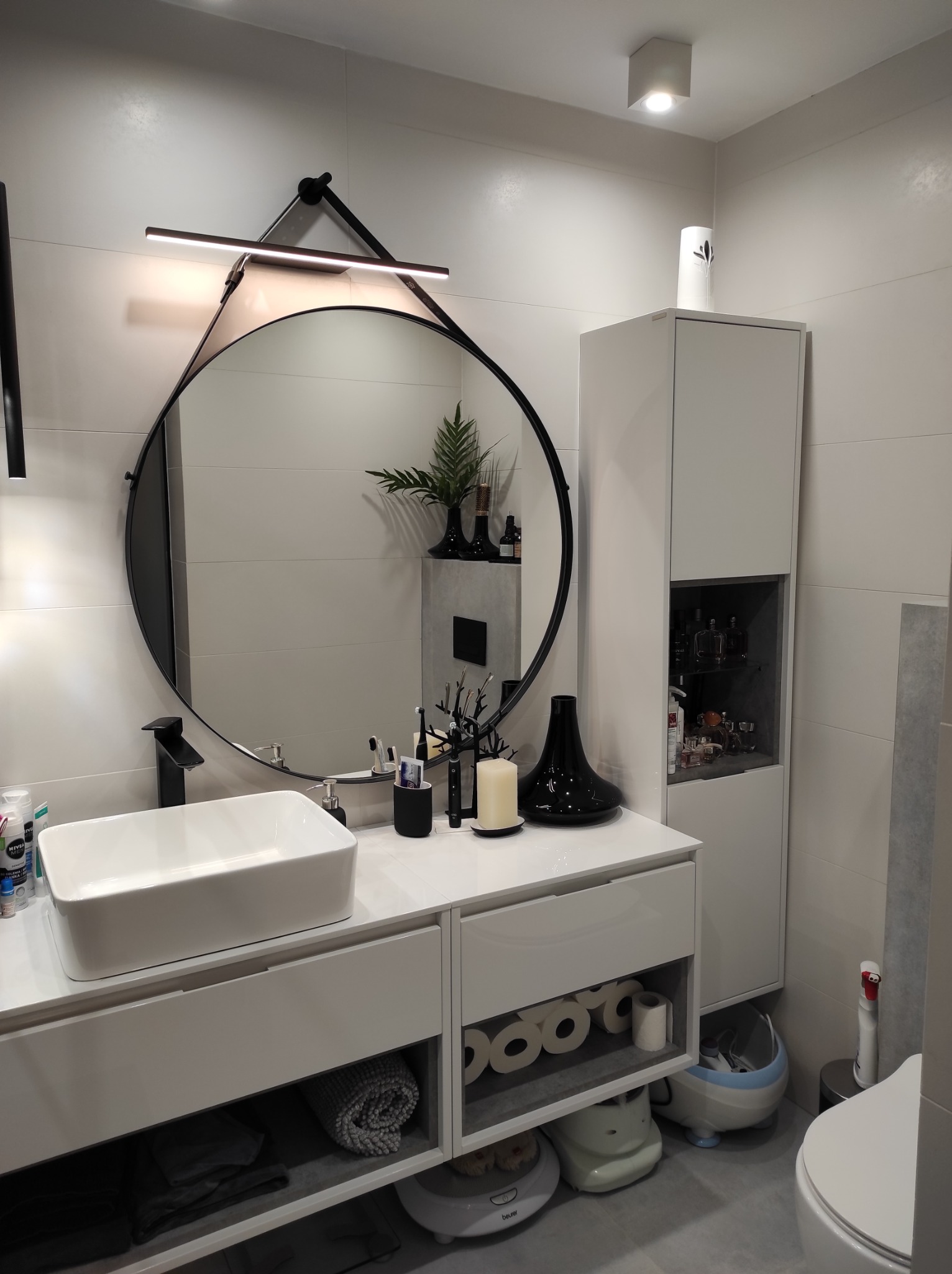 Nowoczesna, jasna łazienka z okrągłym lustrem z podświetleniem LED, białą umywalką nablatową na szafce z szufladami, wysoką szafką z półkami i dekoracjami, oraz elementami wyposażenia jak wazon...