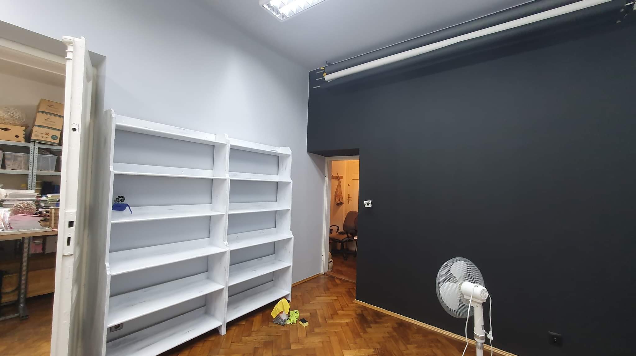 Pokój po odświeżeniu, z pomalowaną na ciemny kolor ścianą, nowymi białymi regałami i wentylatorem pod ścianą, na podłodze parkiet i ściereczki do sprzątania.