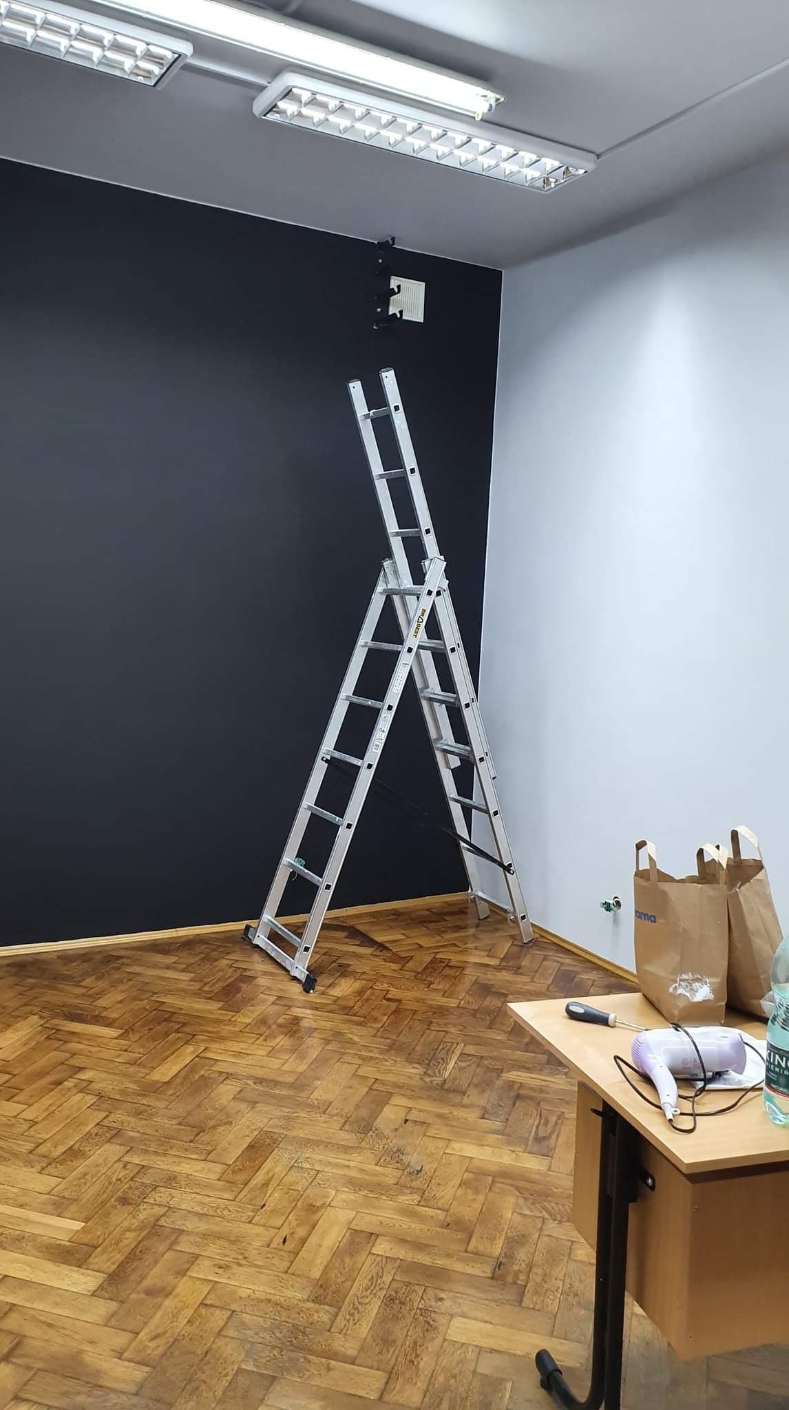 Pokój biurowy z pomalowaną na czarno ścianą, aluminiowa drabina rozstawiona na parkiecie, na biurku suszarka, śrubokręt i butelka wody.