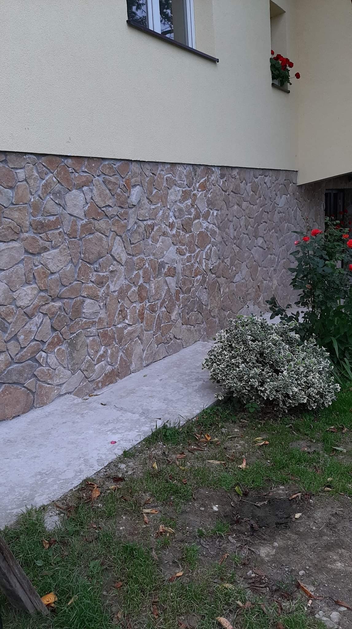 Elewacja budynku z dekoracyjnym wykończeniem z kamienia, widoczne kwiaty w oknie i krzew na trawniku.