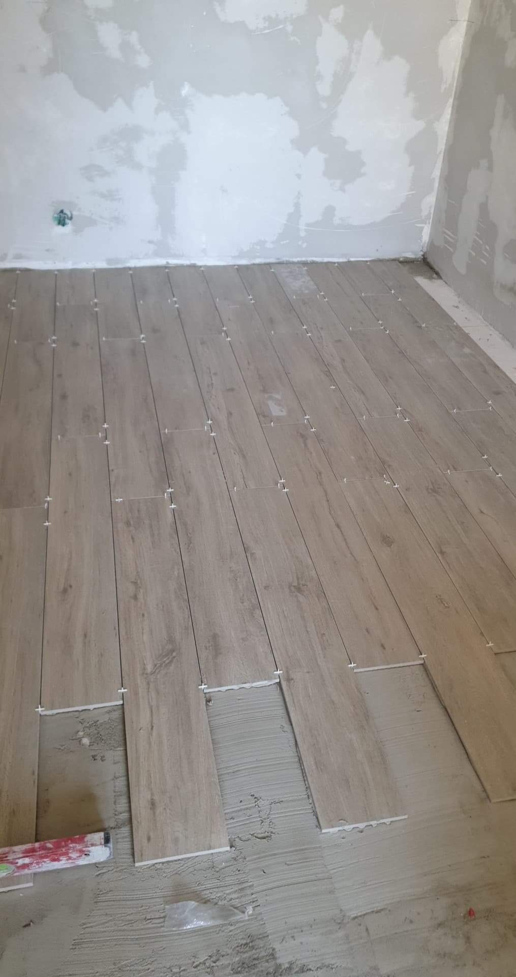 Układanie podłogi z płytek imitujących drewno, widoczne krzyżyki dystansowe i poziomica, surowe ściany w tle.