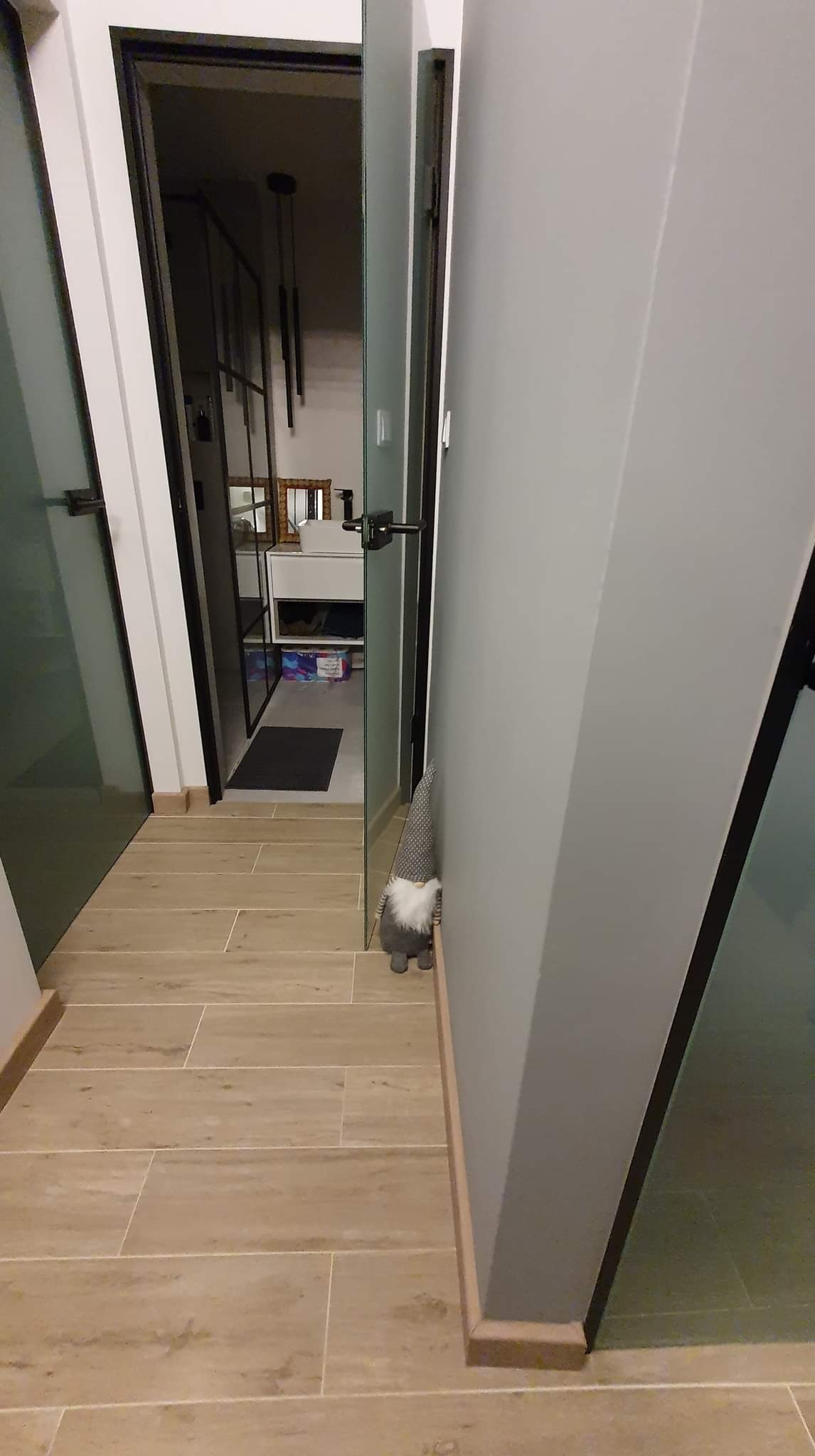 Korytarz z płytkami imitującymi drewno, szarą ścianą i szklanymi drzwiami, z widoczną dekoracją w postaci krasnala przy ścianie.