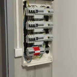 Otwarta, biała skrzynka z zamontowanymi zabezpieczeniami i licznikami energii elektrycznej, widoczne przewody i elementy instalacji, zamontowana na szarej ścianie.