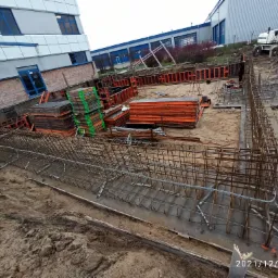 Budowa fundamentów budynku przemysłowego: zbrojenie stalowe, szalunki pomarańczowe i zielone, wykop ziemny, w tle budynek z niebieskimi oknami i koparka.