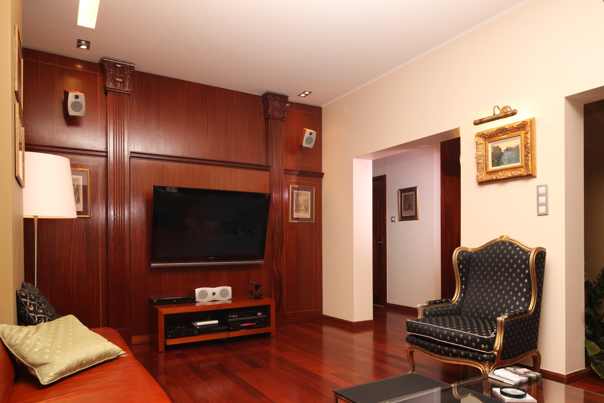 Elegancki salon z drewnianą boazerią, telewizorem, skórzaną sofą i fotelem w stylu Ludwika, oświetlony lampą podłogową i punktowymi reflektorami, z parkietem z ciemnego drewna.