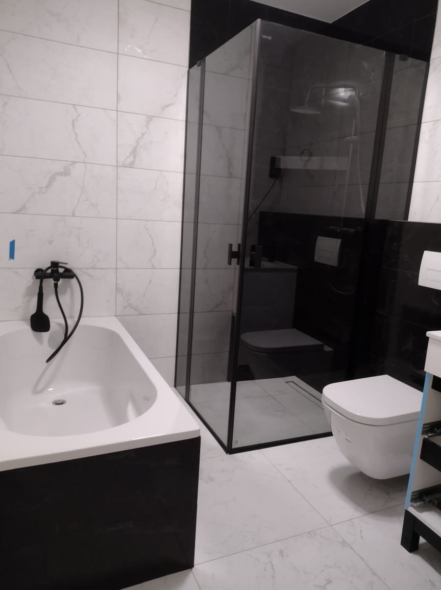 Nowoczesna łazienka z biało-czarnymi płytkami imitującymi marmur, kabina prysznicowa typu walk-in z czarnymi profilami, biała wanna z czarną obudową i czarną armaturą, wisząca toaleta.